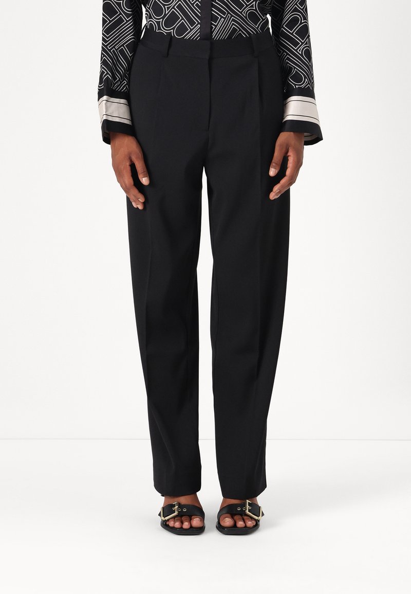 Pantalons noirs taille haute avec une coupe ajustée, présentant des plis à l'avant et des poches latérales. Associés à des sandales noires plates avec des accents dorés.
