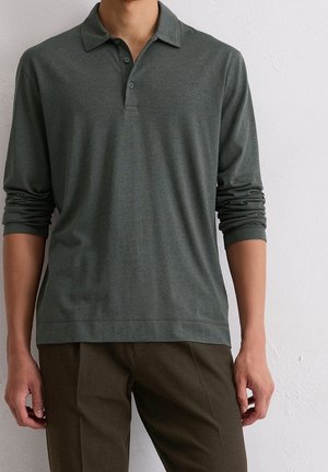 Homme portant un polo gris foncé à manches longues avec des boutons et un pantalon marron foncé, debout contre un mur blanc texturé.