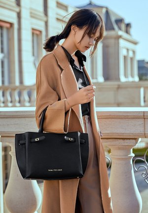 Bolso tote de cuero negro con forma estructurada y detalles metálicos en tono dorado. El bolso cuenta con dos asas y una textura suave, contrastando con un abrigo de color claro.