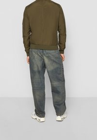 Olivově zelená bomber bunda s elastickými manžetami, kombinovaná s volnými, vybledlými modrými džínami a bílými teniskami.