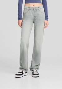 Los pantalones vaqueros rectos de color gris claro cuentan con cinco bolsillos, una textura deslavada y un cierre de botón, combinados con zapatillas negras y blancas.