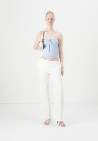 Top fără bretele de culoare albastru deschis, cu un nod în față, asortat cu pantaloni albi cu talie largă. Modelul ține o geantă de umăr crem, stând pe un fundal deschis.