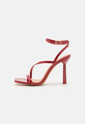 Rød højhælet sandal med tynde stropper, en ankelrem og firkantet åben tå på en ens hvid baggrund.
