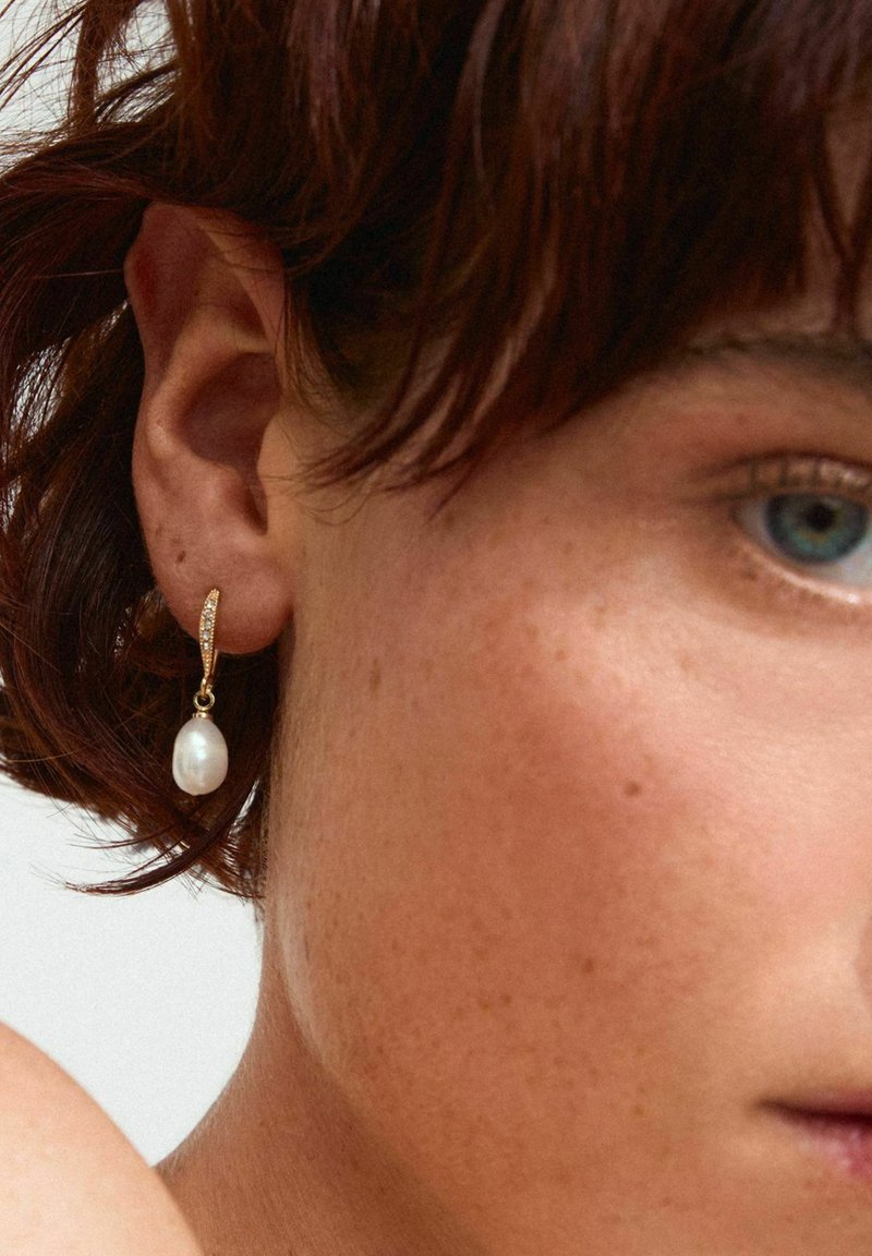 Boucle d'oreille en or avec une perle blanche suspendue et de petites pierres transparentes. Gros plan mettant en avant les détails de l'oreille et de la boucle d'oreille.