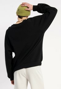 Czarna bluza z luźnym krojem, okrągłym dekoltem i opadającymi ramionami. Modelka nosi zieloną dzianinową czapkę. Białe spodnie widoczne poniżej.