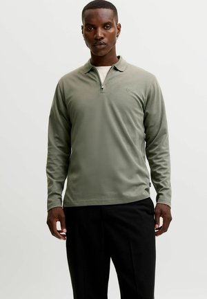 Homme portant un polo vert clair à manches longues avec fermeture éclair sur la moitié et un pantalon noir, debout devant un fond blanc uni.