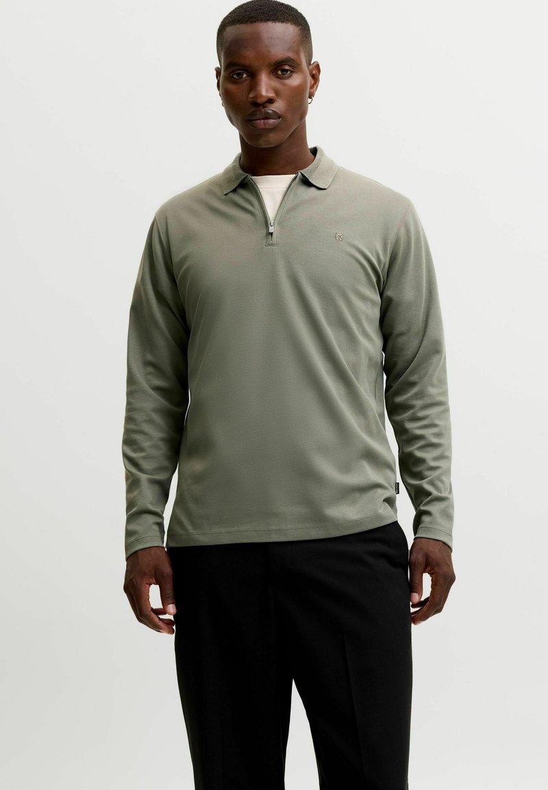 Homme portant un polo vert clair à manches longues avec fermeture éclair sur la moitié et un pantalon noir, debout devant un fond blanc uni.