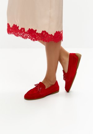 Benen in een beige rok met rode kanten rand en rode suède loafers met bruine zolen, één voet opgetild met de hiel van de vloer.