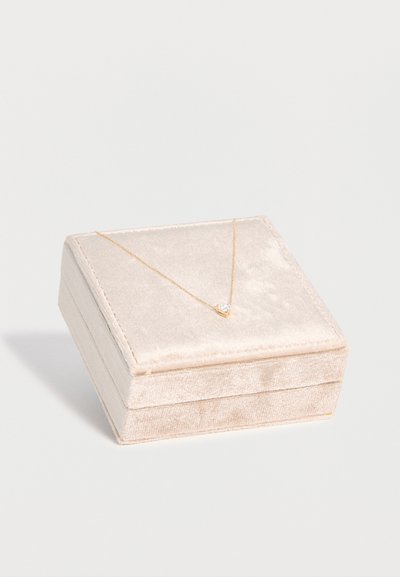 Collier en or délicat avec un petit pendentif en pierre précieuse transparente reposant sur un écrin à bijoux en velours beige doux sur fond blanc.