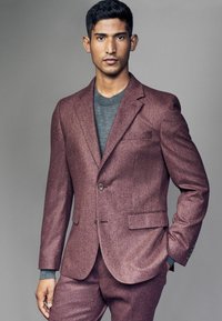 Costume en laine bordeaux à coupe ajustée, avec un col cranté, un bouton unique et des poches latérales. Le tissu texturé contraste avec un pull gris.