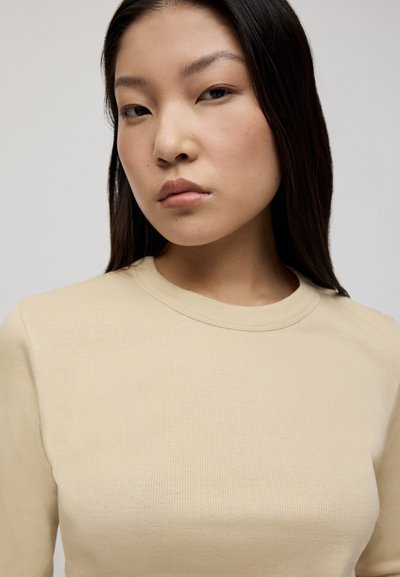 Beige geripptes Langarmoberteil mit rundem Halsausschnitt. Der Stoff hat eine glatte Textur und sitzt eng am Körper, wodurch eine klare Silhouette betont wird.