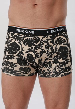 Άνδρας που φοράει μπεζ μποξεράκια με μαύρο τροπικό φύλλωμα και μαύρη μέση με την επωνυμία "Pier One".