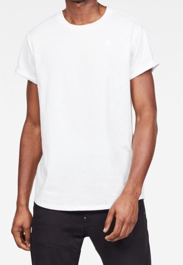 Homme portant un t-shirt blanc uni à manches courtes et col rond et un pantalon noir, debout devant un fond gris clair uni.
