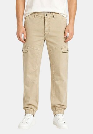 Uomo con pantaloni cargo beige con polsini elasticizzati e scarpe da ginnastica bianche, in piedi con le mani rilassate lungo i fianchi.