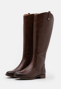 Une paire de bottes en cuir marron foncé, hautes, avec des orteils arrondis, de petits talons et des fermetures éclair latérales, se tenant debout sur un fond blanc.