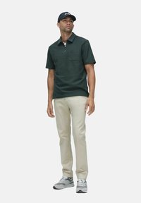 Mörkgrön kortärmad polo med en framficka, kombinerad med beige byxor och grå sneakers med vita detaljer.