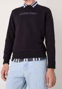 Felpa nera con collo tondo e logo "Calvin Klein". Indossata sopra una camicia a righe blu e bianche, abbinata a jeans azzurri chiari.