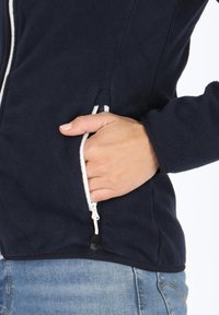Navy fleece-takki valkoisilla vetoketjuyksityiskohdilla; sisältää vetoketjullisen taskun sivussa ja teksturoitu materiaali.