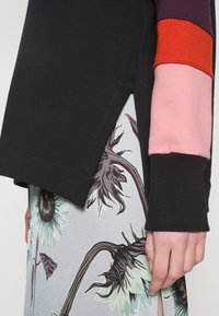 Felpa nera con maniche multicolori (rosso, rosa, viola) e fessure laterali, abbinata a una gonna con motivo floreale caratterizzata da girasoli.