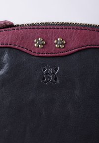 Black leather bag with a maroon top panel, decorative brass flower accents, embossed logo, and a gold zipper along the top edge.