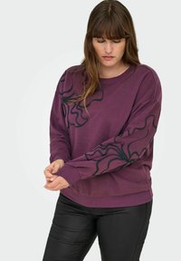 Sudadera morada con gráficos de líneas onduladas negras en las mangas, cuello redondo, mangas largas y puños acanalados. Combinada con pantalones de cuero negros.