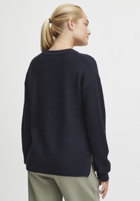 Pull en maille bleu marine avec un motif texturé, coupe décontractée, col rond et poignets côtelés. La vue de dos présente de légentes fentes sur les côtés.