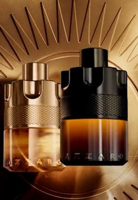 Deux flacons de parfum Azzaro, un doré et un noir avec de l'ambre, placés sur un fond doré texturé.