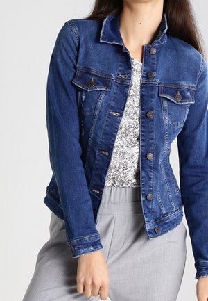 Femme portant une veste en jean bleue sur un haut blanc à fleurs et un pantalon gris clair, posant avec une main près de la hanche.