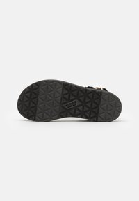 Suela de sandalia de goma negra que presenta un patrón texturizado con formas geométricas y el logo de Teva. Diseñada para ofrecer tracción y durabilidad.