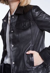 Giubbotto di pelle nera con chiusure a pressione, due tasche frontali, colletto classico e texture liscia con dettagli di cucitura verticale.