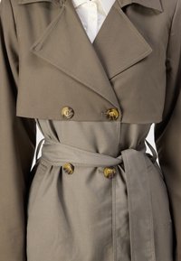 Kaki trenchcoat med doble knapper, brede revers, matchende belte og glatt stofftekstur. Har uttalte sømdetaljer.