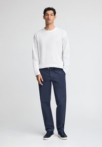 Vit långärmad skjorta med ett strukturerat mönster, marinblå byxor och mörka sneakers med vit sula. Modellen står mot en ljus bakgrund.