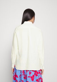 Camicia di lino giallo chiaro con maniche lunghe, una leggera lucentezza, polsini con bottoni e una piega sulla schiena, abbinata a una gonna blu con motivo floreale.