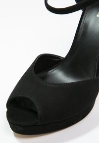 Chaussure à talon haut en daim noir à bout ouvert avec bride à la cheville et semelle plateforme sur fond blanc.