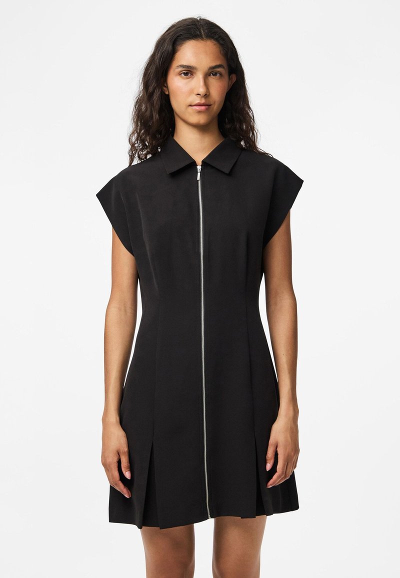 Vestido preto com um design curto e estruturado, apresentando um fecho de correr à frente e mangas curtas. Tecido suave, caimento ajustado e saia plissada.