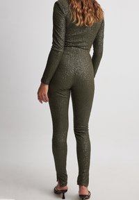 Olivgrön jumpsuit med långa ärmar, ribbad textur och subtila glitterdetaljer, figurnära passform och ankellängd. Bakifrån visad.