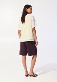 Top senza maniche giallo chiaro sopra una camicia bianca, abbinato a pantaloni corti viola scuro e sandali con tacco marroni, mostrato di schiena.