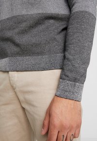 Harmaa raidoitettu neulepaita, jossa on ribbaatut hihansuut ja helma. Kangas näyttää sileältä ja se on yhdistetty vaaleanbeigeihin housuihin. Käsi, jossa on näkyvissä tatuointi.