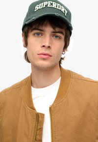 Chaqueta bomber caqui con cuello de canalé, camiseta blanca por debajo, gorra verde con el logotipo "SUPERDRY" en blanco, modelo con pelo de longitud media.
