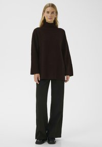 Pull à col roulé côtelé marron avec des manches longues, silhouette ajustée, associé à un pantalon ample noir et des chaussures noires massives.