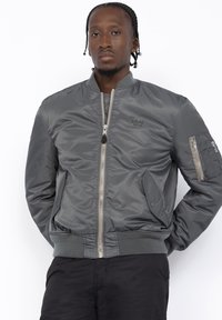 Giacca bomber grigia con chiusura zip, polsini e orlo a coste, due tasche laterali e una tasca aggiuntiva sul braccio. Texture liscia.