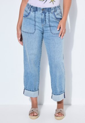 Kvinde iført lyseblå smalle denimjeans med elastisk taljebånd og forlommer, kombineret med beige åbne kilehælssandaler.