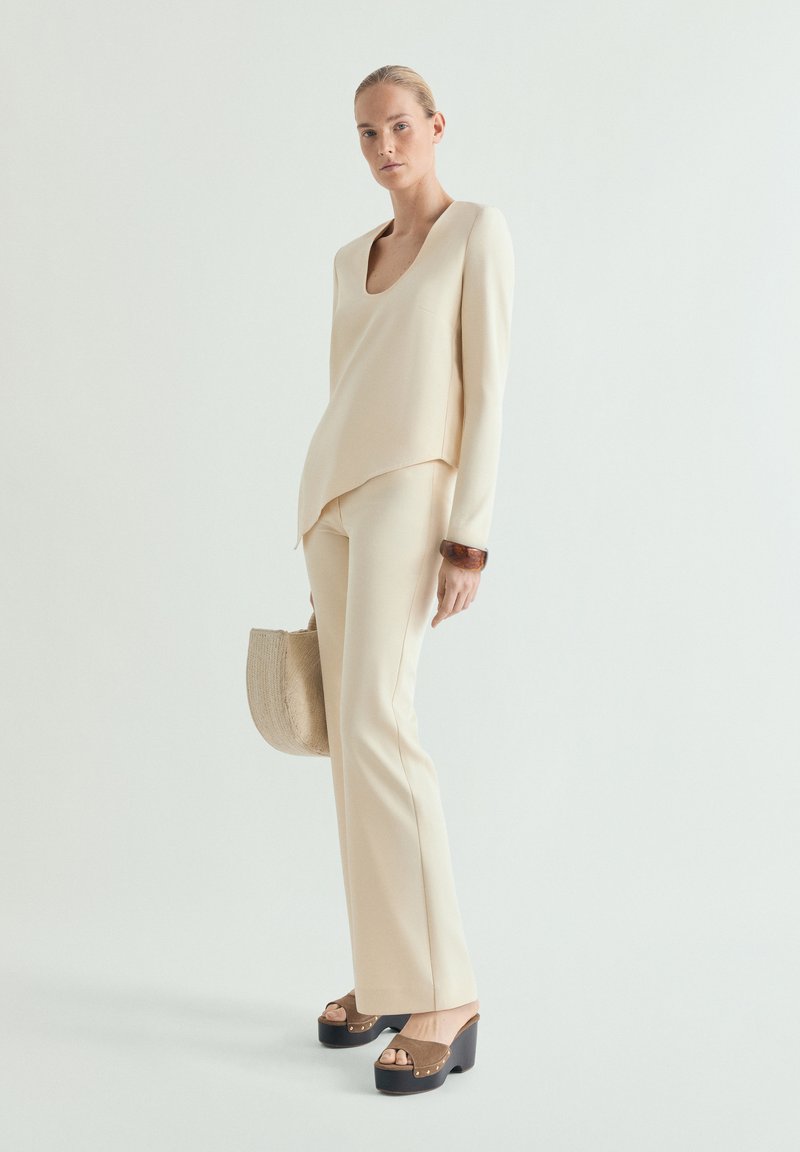 Mujer con top y pantalones color crema, sosteniendo un bolso tejido beige, y calzando sandalias de plataforma marrones, posando frente a un fondo neutro.