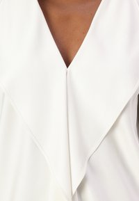 Blusa bianca fluente con scollatura a V profonda, caratterizzata da un design del colletto a strati e una texture liscia. Cuciture ordinate lungo i bordi.