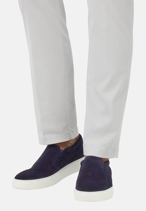 Chaussures slip-on en suède bleu marine avec une semelle en caoutchouc blanche, présentant une texture lisse et des panneaux latéraux élastiques. Associées à un pantalon gris clair.