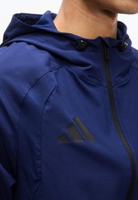 Κοντινό πλάνο ατόμου που φοράει μπλε σκούρο μπλούζα με φερμουάρ και κουκούλα, με μαύρο λογότυπο τριών ρίγων στο αριστερό στήθος.