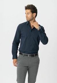 Mand justerer kraven, iført en marineblå skjorte med knapper, grå bukser, et sort bælte og et sølvfarvet armbåndsur mod en ensfarvet hvid baggrund.