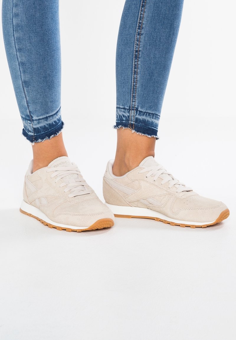 Personne portant des baskets en daim beige clair avec des lacets blancs et des semelles en gomme, associées à un jean denim bleu aux ourlets effilochés.