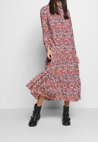 Abito midi floreale con maniche lunghe, caratterizzato da uno sfondo rosa e fiori multicolori, vita cintrata e orlo a strati con rouches. Indossato con stivali neri.