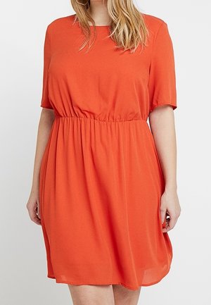 Donna che indossa un vestito arancione a maniche corte, lunghezza al ginocchio, con una cintura elastica in vita e scollo rotondo, in piedi contro uno sfondo semplice.